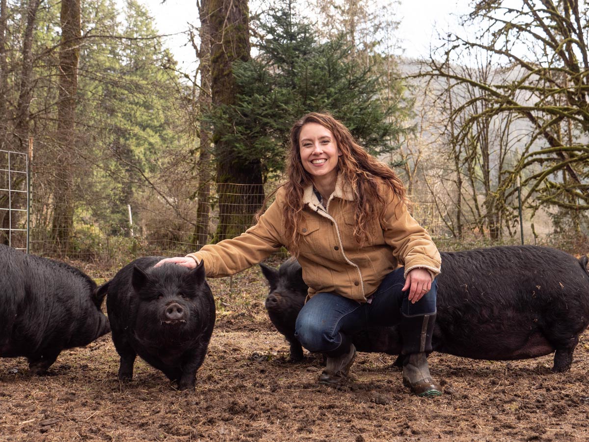 A woman crouched down next to four American Guinea Hogs out in the pasture.