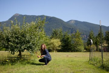 A woman crouched down next to an established fruit tree with mountains in the background.