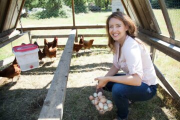 A woman crouched down inside a portable chicken tractor holding a basket full of eggs.