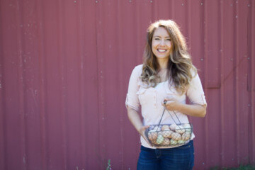 A woman standing in front of a red barn holding up a wire basket full of farm fresh eggs.