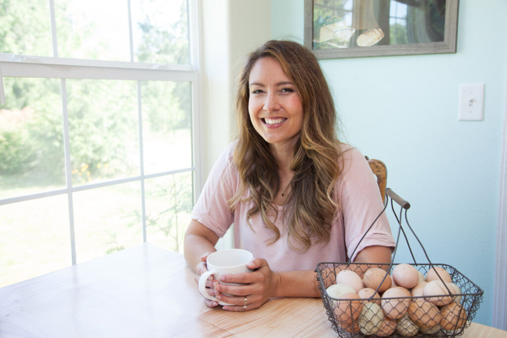 A woman sitting at her kitchen table with a cup of coffee next to a wire basket filled with farm fresh eggs.