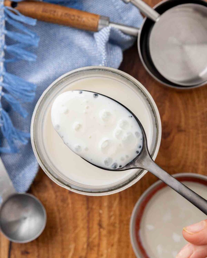 A bird's eye view of a jar of homemade buttermilk with a hand lifting a spoonful to show texture.