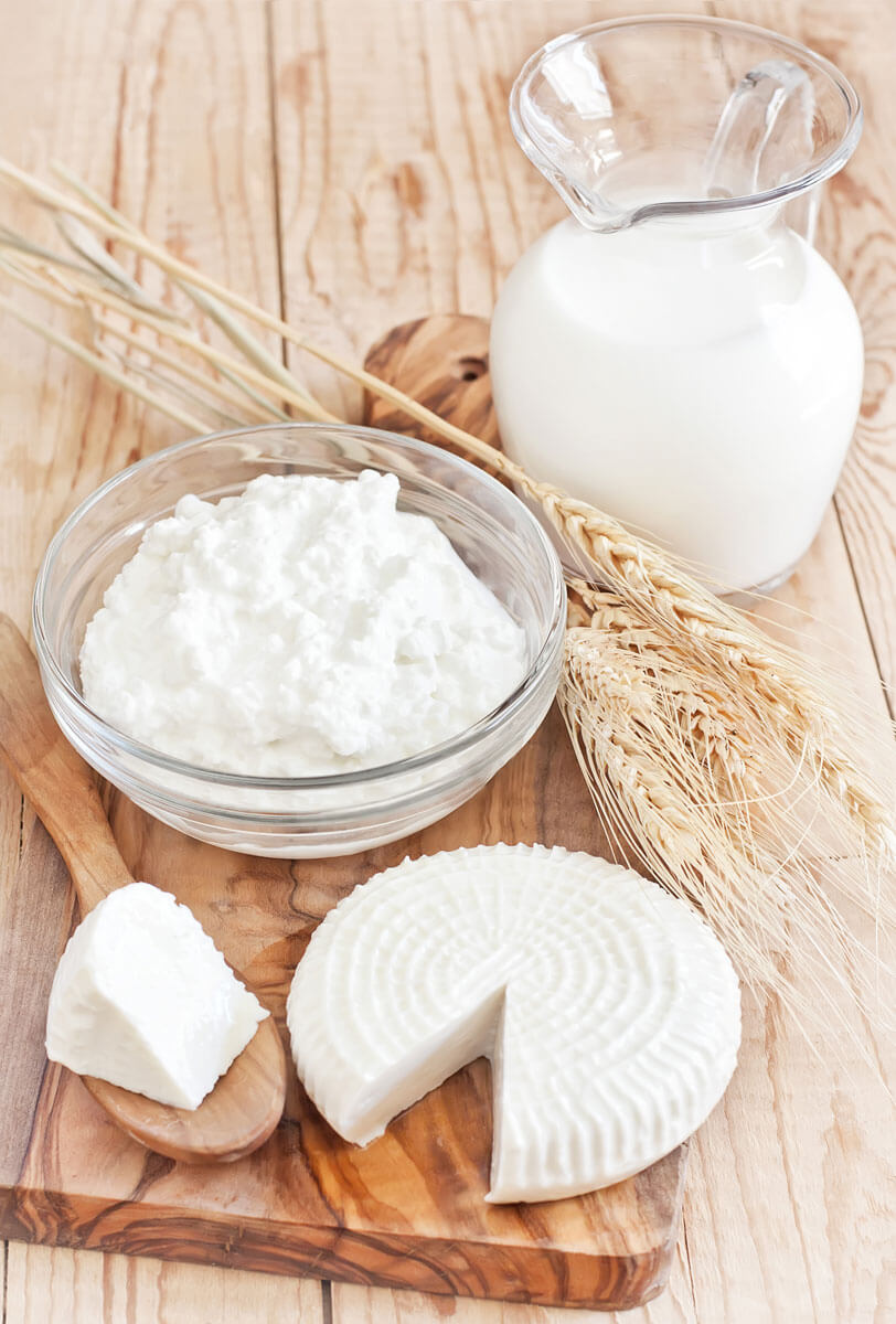 A wheel of soft cheese on a wooden cutting board next to a glass bowl of thick greek yogurt and a glass pitcher of milk in the background.