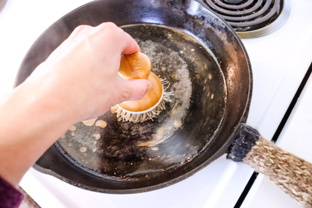 A hand scrubbing at the bottom of a cast iron skillet on the stovetop.