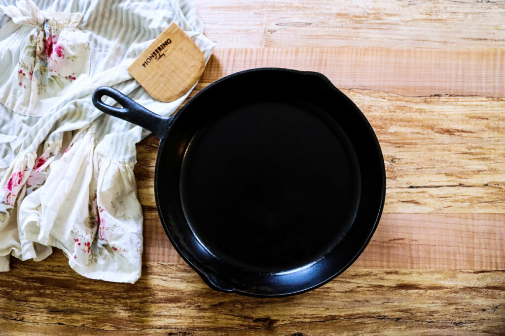 A clean cast iron pan sitting on a wooden counter top with a towel and a wooden bench scraper beside it.