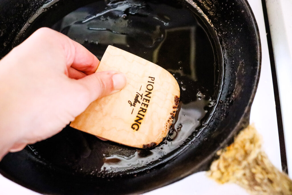 A cast iron pan being cleaned with a wooden bench scraper.