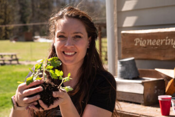 A woman holding seedlings in her hands, standing outside by a potting bench.