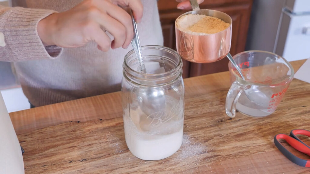 Image of a hand scooping in flour into a mason jar with sourdough starter.