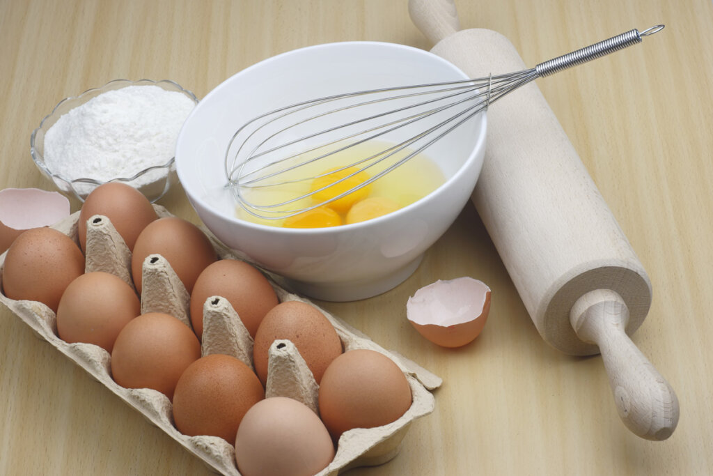 Eggs cracked into a white bowl with egg carton, rolling pin and wire whisk beside it.