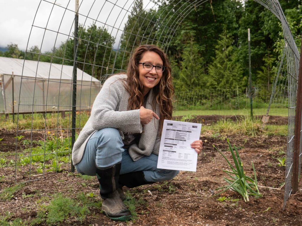A woman crouched in the garden holding up a paper with soil test results.