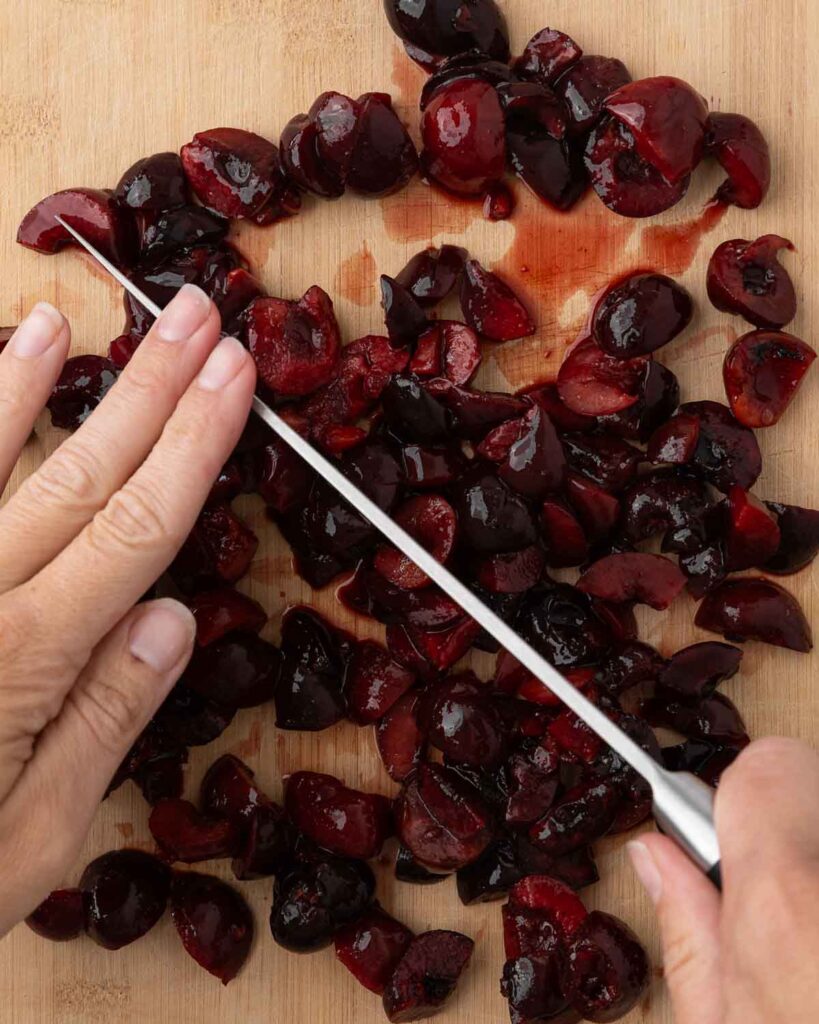 A hand holding a knife chopping cherries on a cutting board.