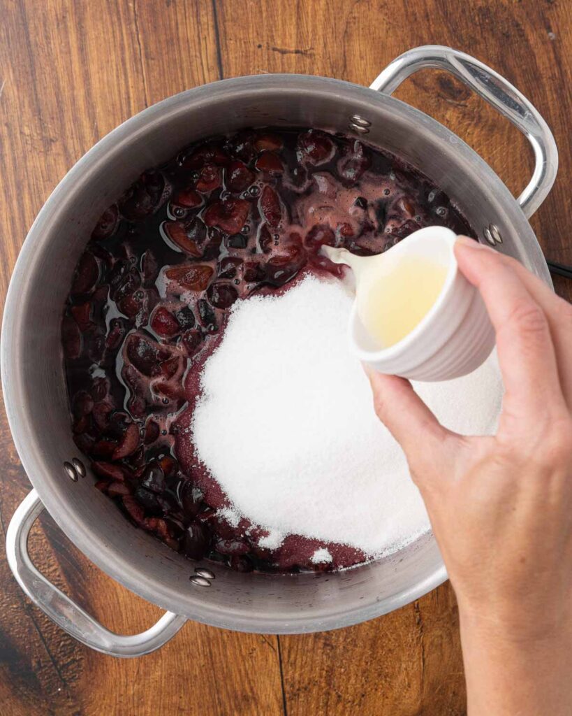 A hand adding lemon juice to a stockpot full of cherries and sugar.