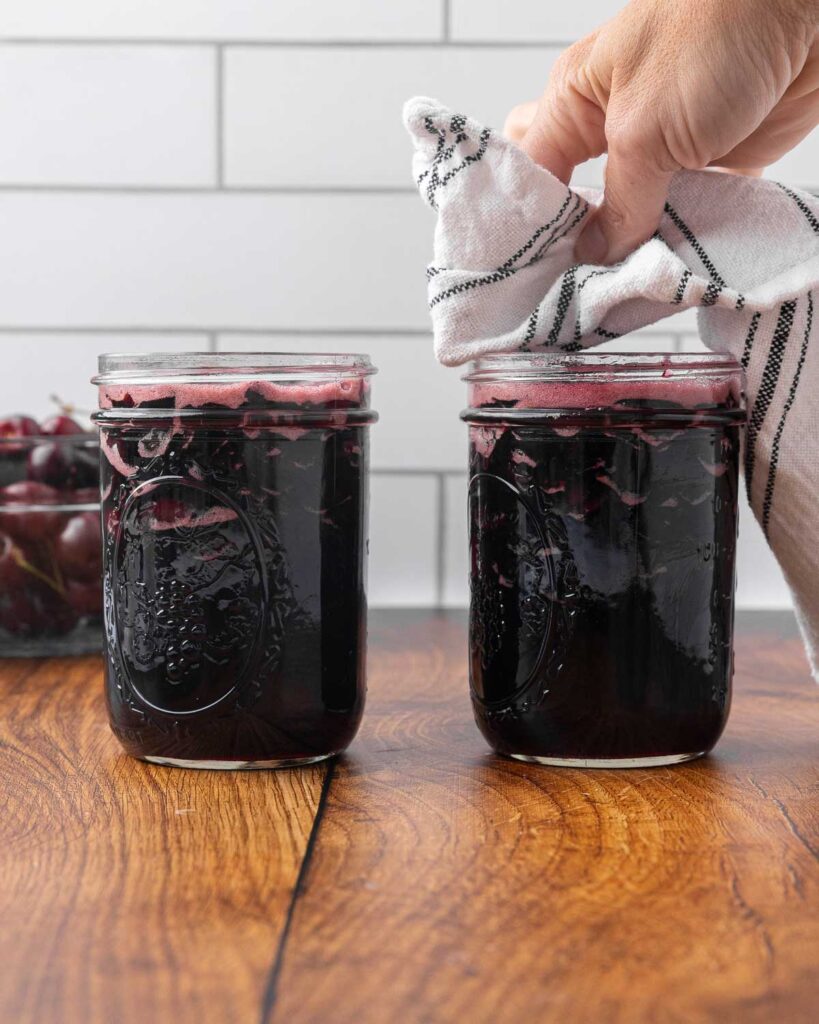 A hand holding a tea towel wiping the rim of a jar full of cherry jam.
