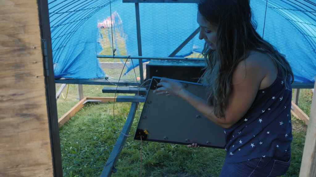 A woman holding up a heat plate inside a mobile chicken coop.