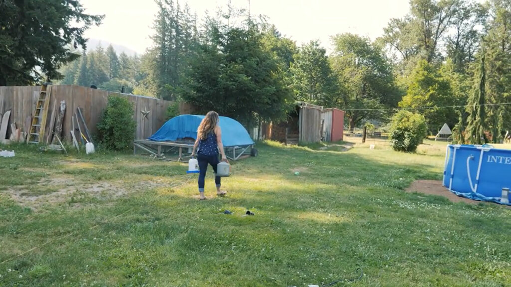 A woman walking through the grass to a mobile chicken tractor carrying two waterers.