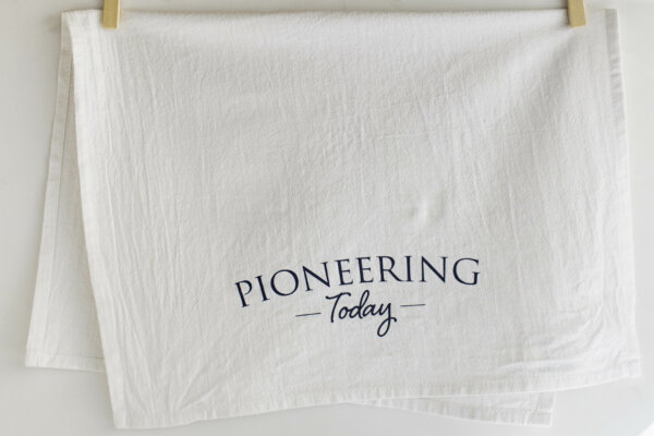 Pioneering Today cotton tea towel handing from wooden clothespins