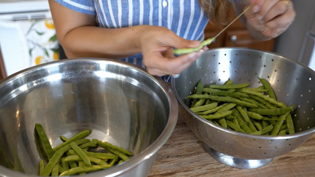 A woman stringing green beans and putting them into a stainless steel bowl.