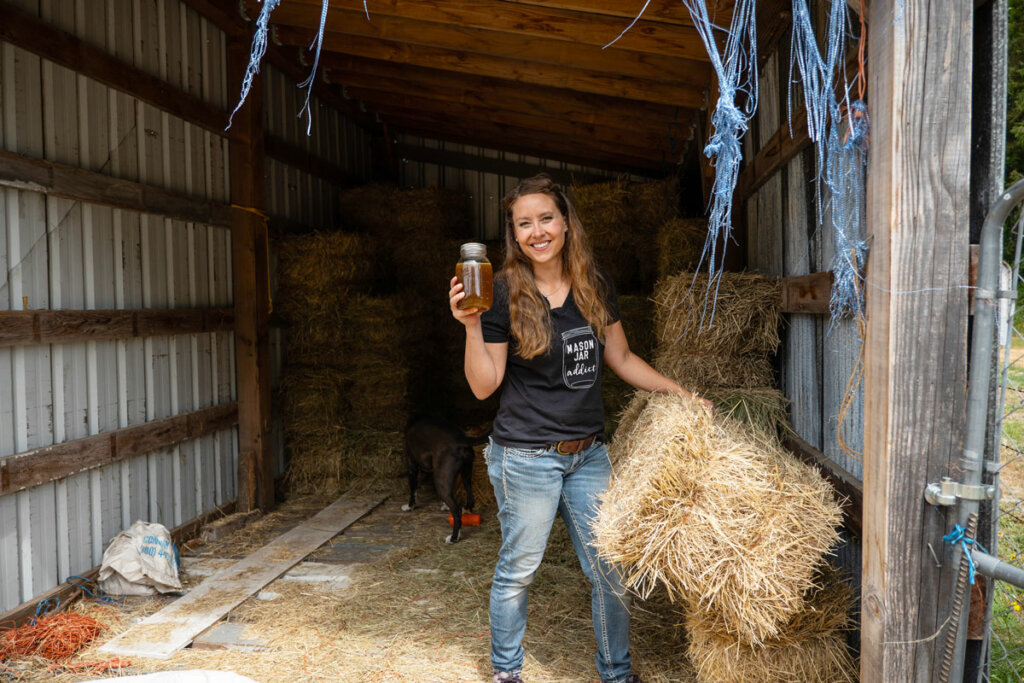 A woman in a hay barn holding up a jar of ginger water.