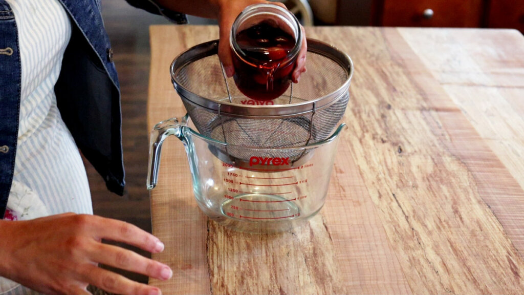 Fruit vinegar being strained through a metal sieve into a glass measuring bowl.