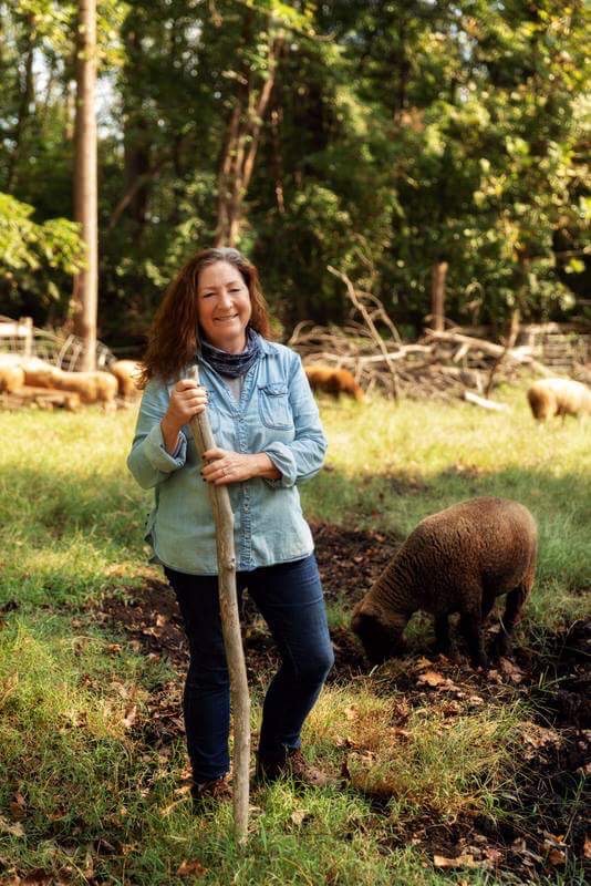 A woman with a walking stick in nature with a sheep in the background.