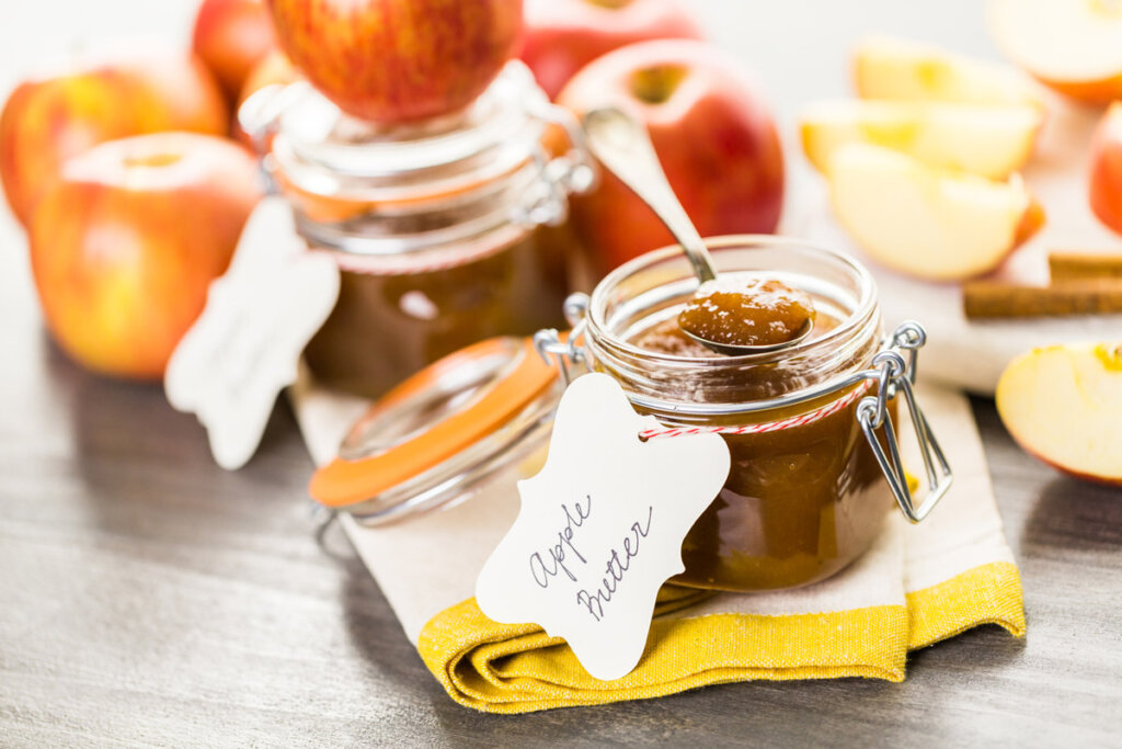 Two jars of homemade apple butter in swing-top jars with tags. Sliced and whole apples and cinnamon sticks surround the jars.