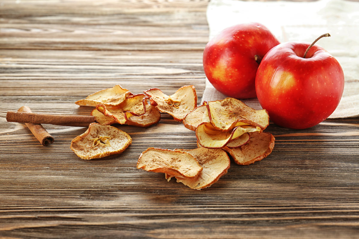 Homemade dehydrated apple chips with cinnamon sticks and two whole apples sitting on a wooden countertop.