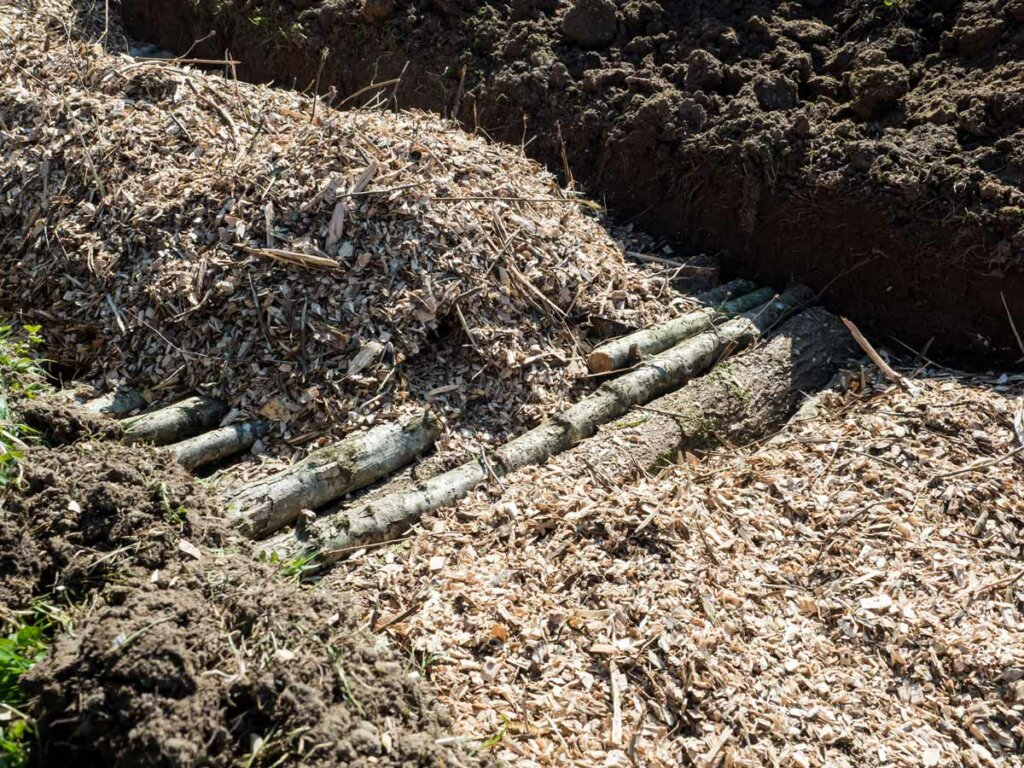 A hugelkultur bed in the ground with layers of logs and wood chips.