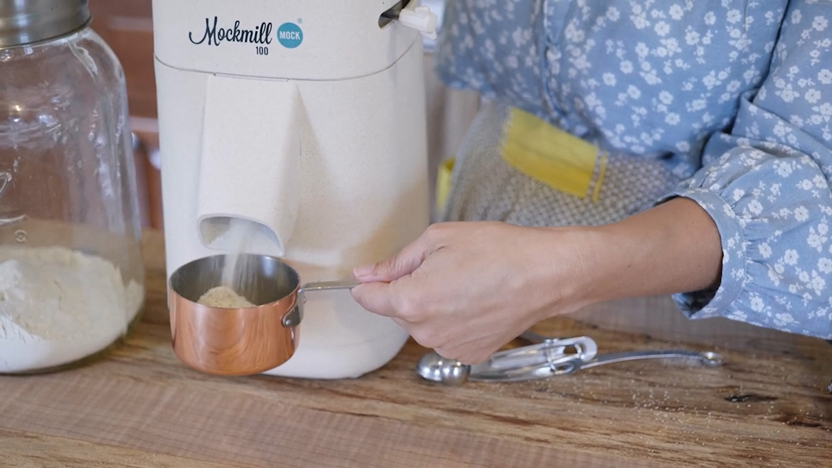 A woman's hands holding a measuring cup catching fresh milled flour from a Mockmill home grain mill.