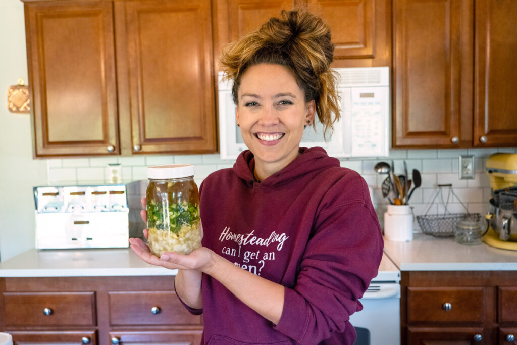 A woman in the kitchen holding a Mason jar filled with fire cider ingredients.