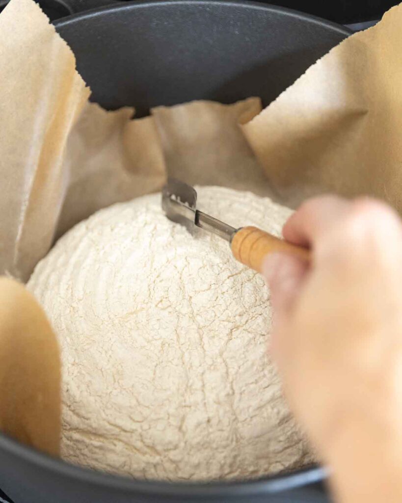 Hands using a bread lame to score a loaf of unbaked no knead artisan bread.