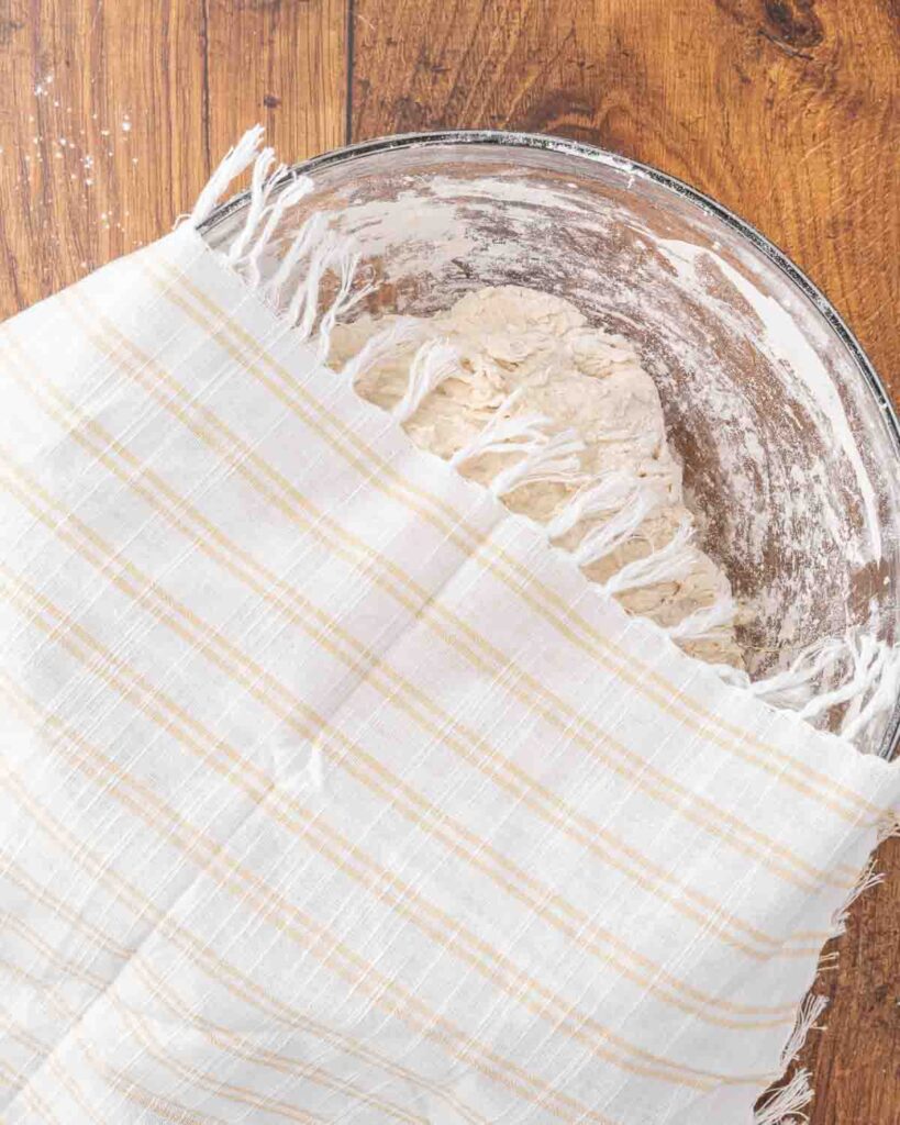 A towel loosely covering a bowl of no knead artisan bread dough.