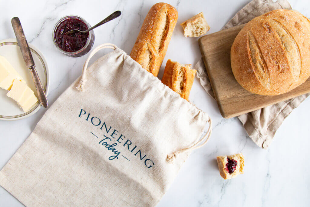 Linen bread bag with two baguettes, cutting board with round loaf of artisan bread, butter and jam on counter top