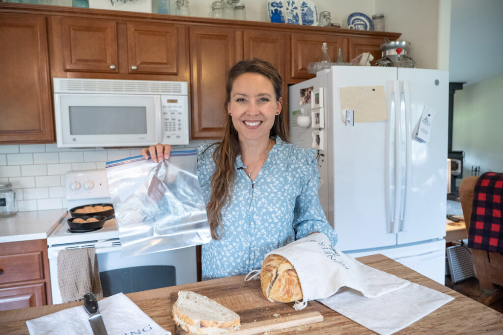 A woman holding up a plastic ziplock bag with a loaf of homemade bread in a linen bread bag on the counter.