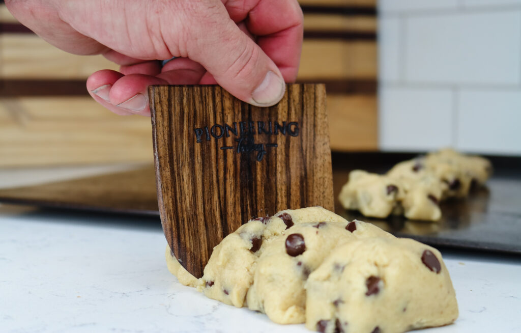 Black walnut wooden dough scraper slicing through log of chocolate chip cookie dough