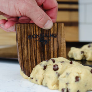 Black walnut wooden dough scraper slicing through log of chocolate chip cookie dough