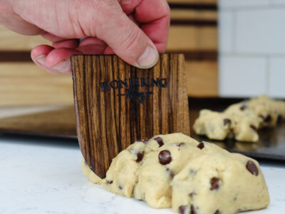 Black walnut wooden dough scraper slicing through log of chocolate chip cookie dough