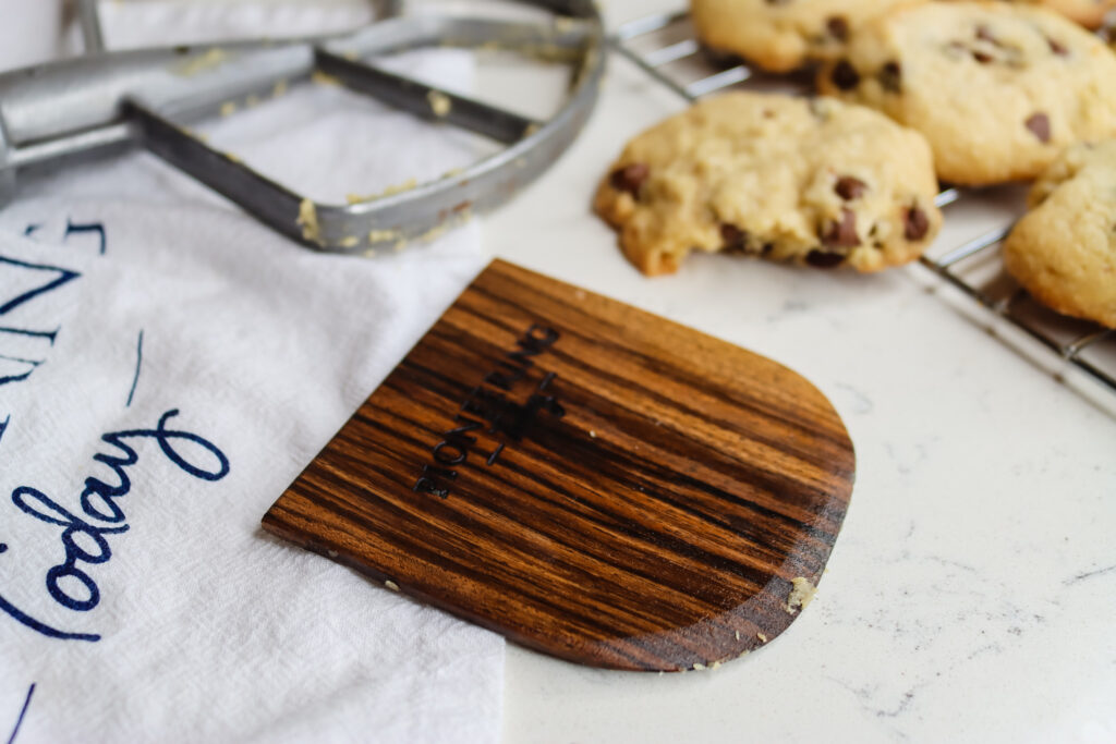 black walnut wooden dough scraper on tea towel with homemade chocolate chip cookies cooling on rack