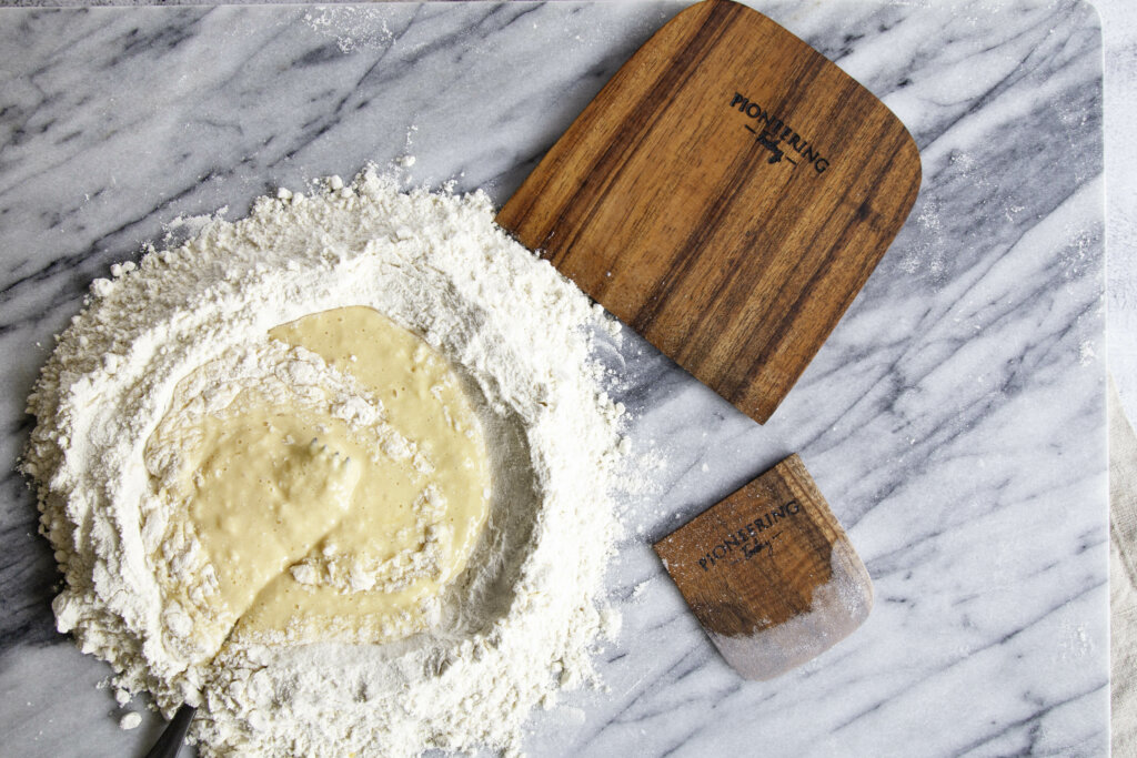 Koa wood bench knife on marble counter top next to flour and egg dough