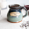 loose leaf tea brewing in handmade pottery mug on counter top with sprinkles of dried herbs on counter and in mason jar