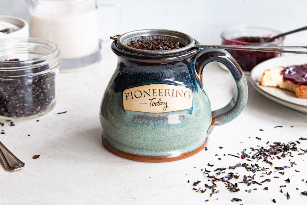 loose leaf tea brewing in handmade pottery mug on counter top with sprinkles of dried herbs on counter and in mason jar