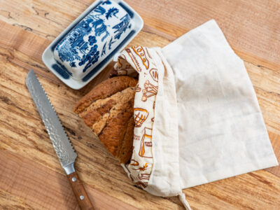 Sliced loaf staying fresh in a USA-made beeswax-lined organic cotton Pioneering Today bread bag on a wood cutting board.