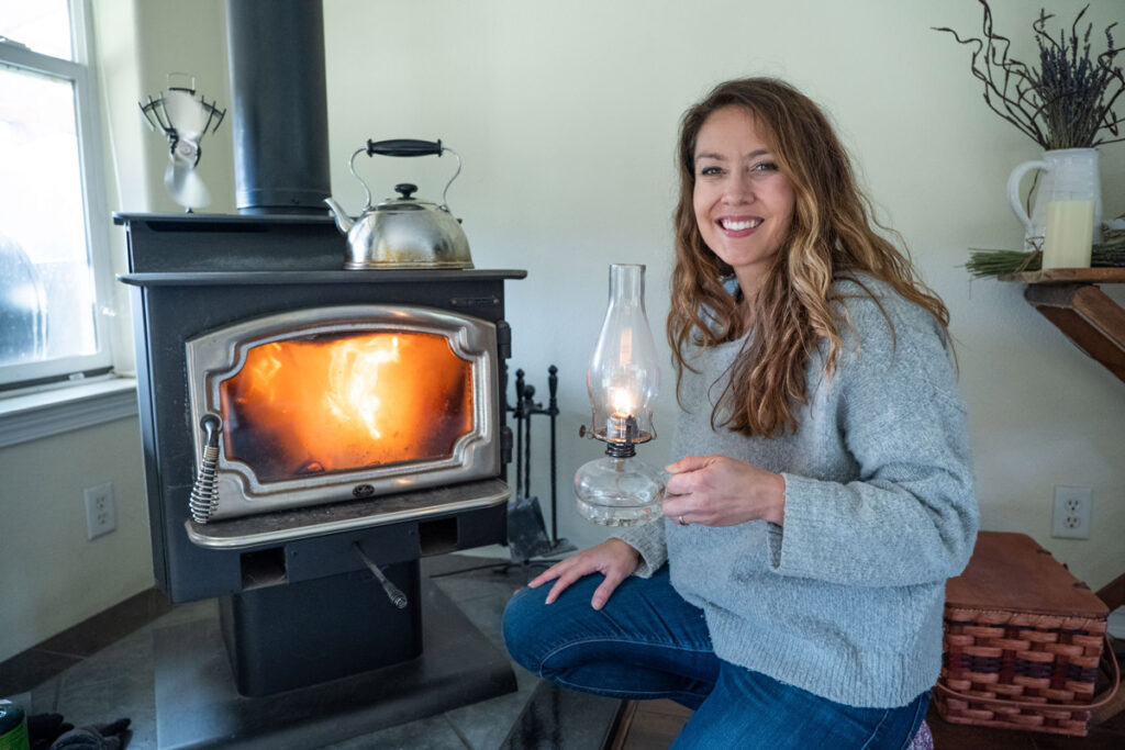 A woman crouched by a woodstove with a fire going, holding an oil lamp. 