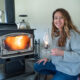 A woman crouched by a woodstove with a fire going, holding an oil lamp.