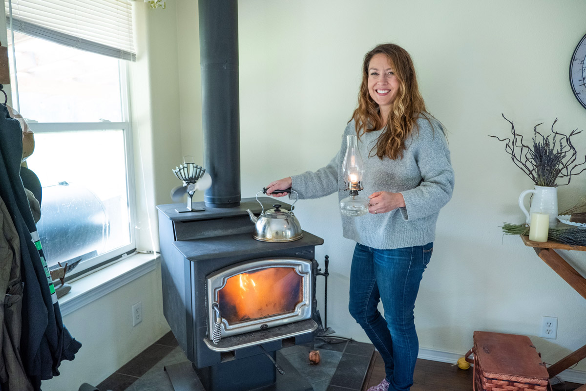 A woman standing by a woodstove with an oil lamp in hand and grabbing a tea kettle.