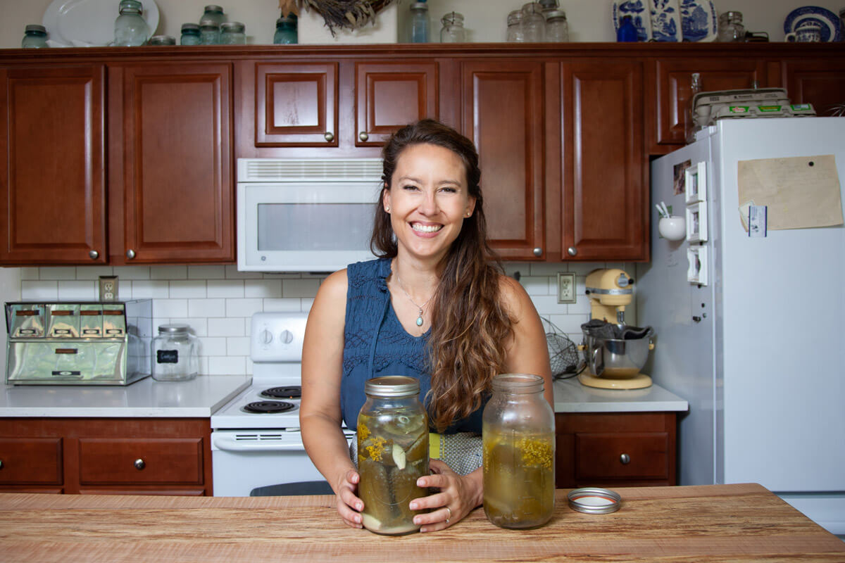 A woman standing behind two large jars of homemade pickles.