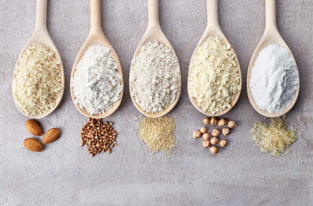 Five wooden spoons with various gluten free flours in them, with the flour source below the spoons.