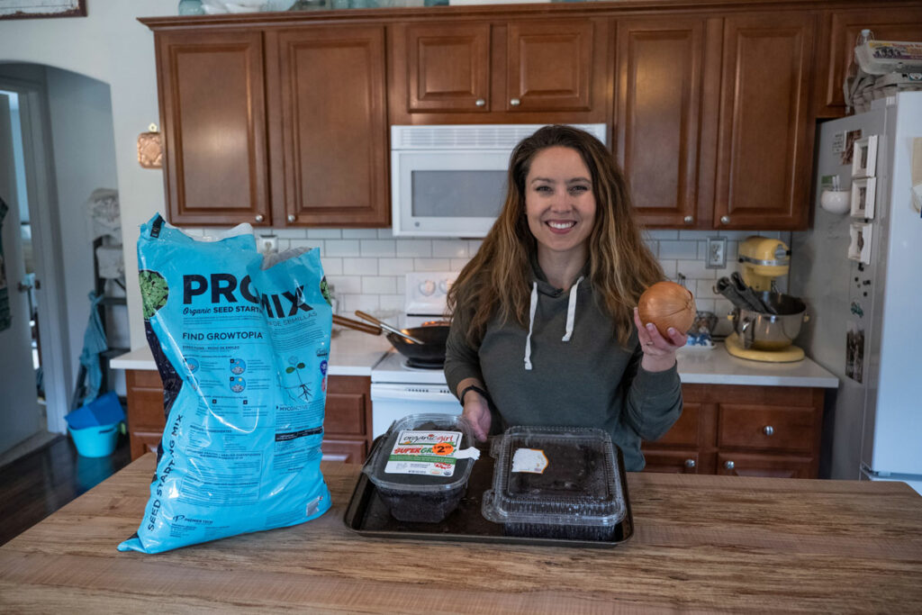 A woman holding an onion with seed starting supplies on the counter in front of her.