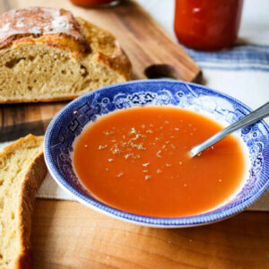 Up close shot of a bowl of tomato soup with a loaf of bread in the background.