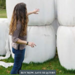 Pinterest pin for saving money on animal feed. A woman standing next to large bales of haylage.