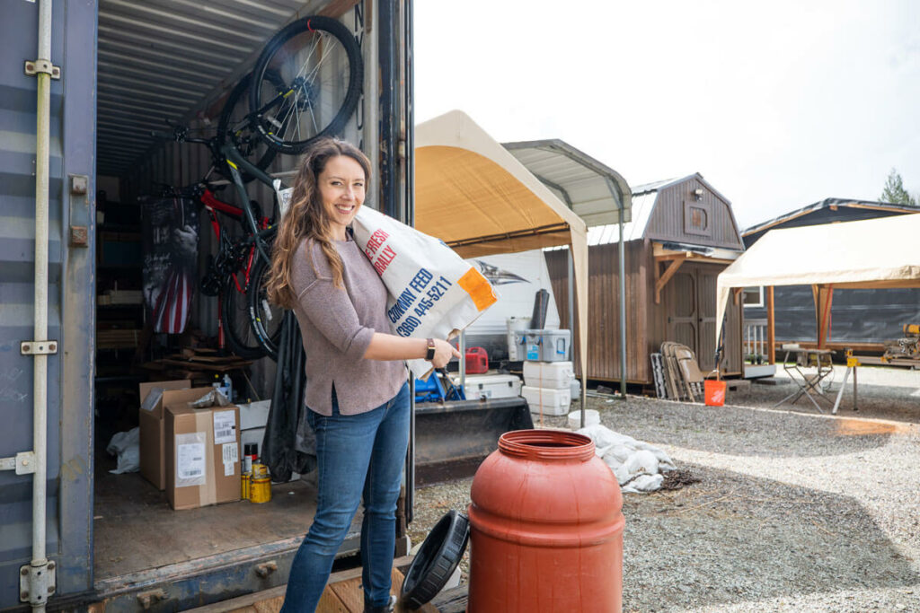 A woman holding a bag of feed by an open 55 gallon drum.